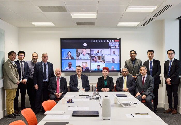 A group of people in a room, standing and sitting in front of a video call on a screen