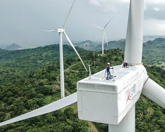 Two workers sit on top of a wind turbine's machine, with other turbines in the background surrounded by green hills.